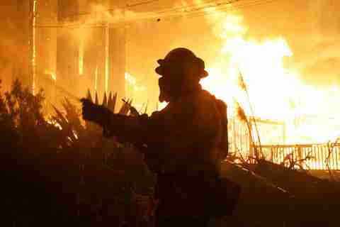 The shadow of a firefighter can be seen in the forefront as a fire rages in the background. 