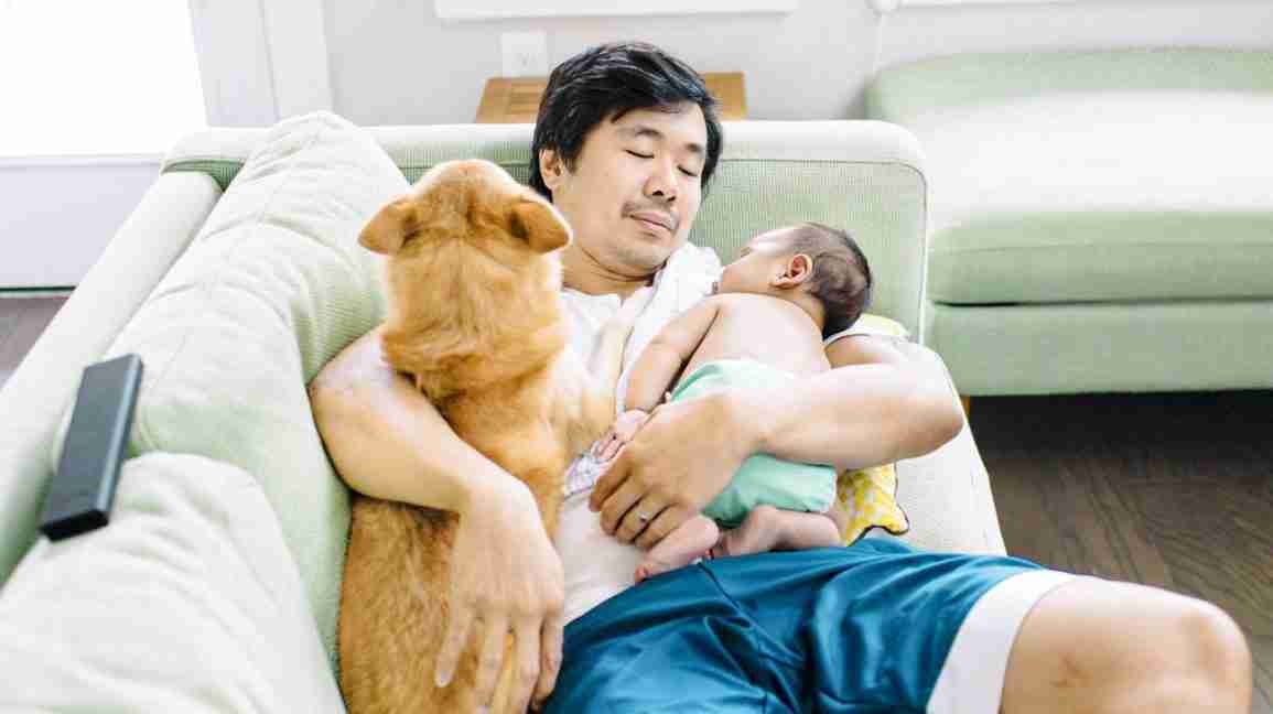 man cuddles on the couch with a dog and a baby