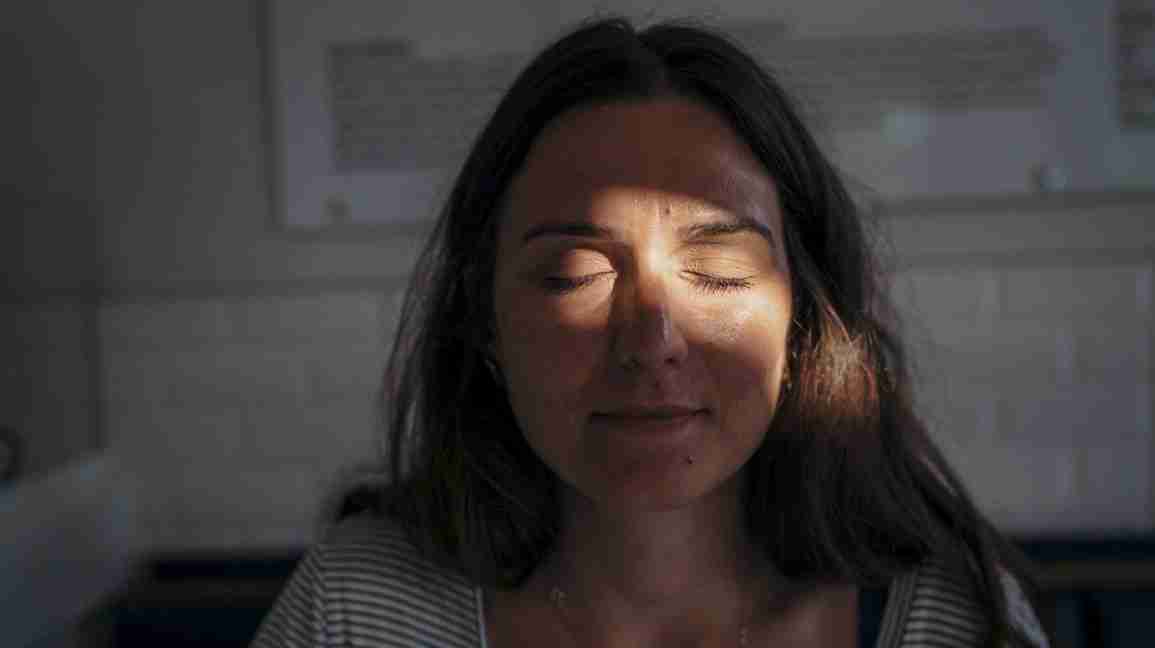 A woman sits in a darkened room with her eyes closed due to a migraine. 