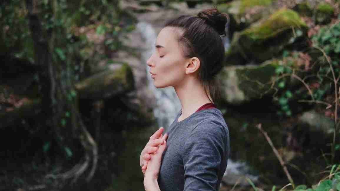 A woman stands in front of a waterfall, with her eyes closed while doing breathing exercises. 