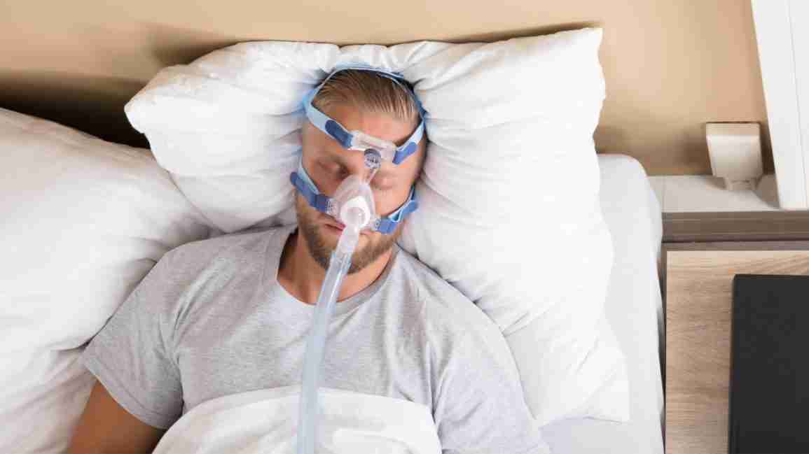 A man lies on his back in bed with a CPAP mask on his face. 