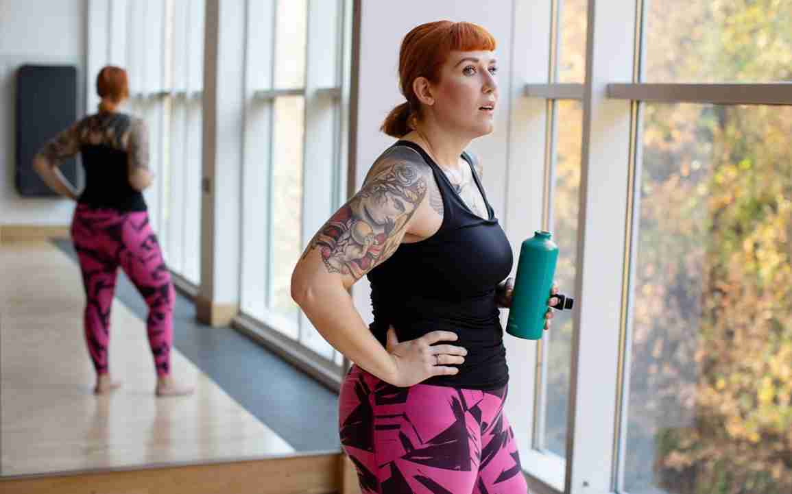 A woman in workout gear holds a water bottle while looking out of a window. 