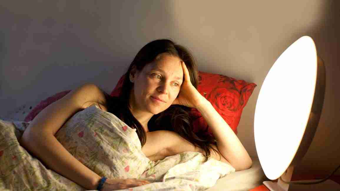 A woman lies  in bed with a light box next to her. 